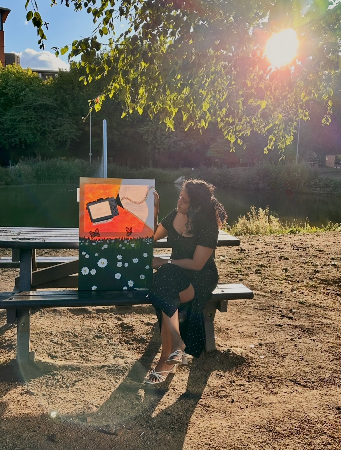 The author sitting on a wooden bench by a canal at golden hour, beside one of her paintings of daisies and a sunset.